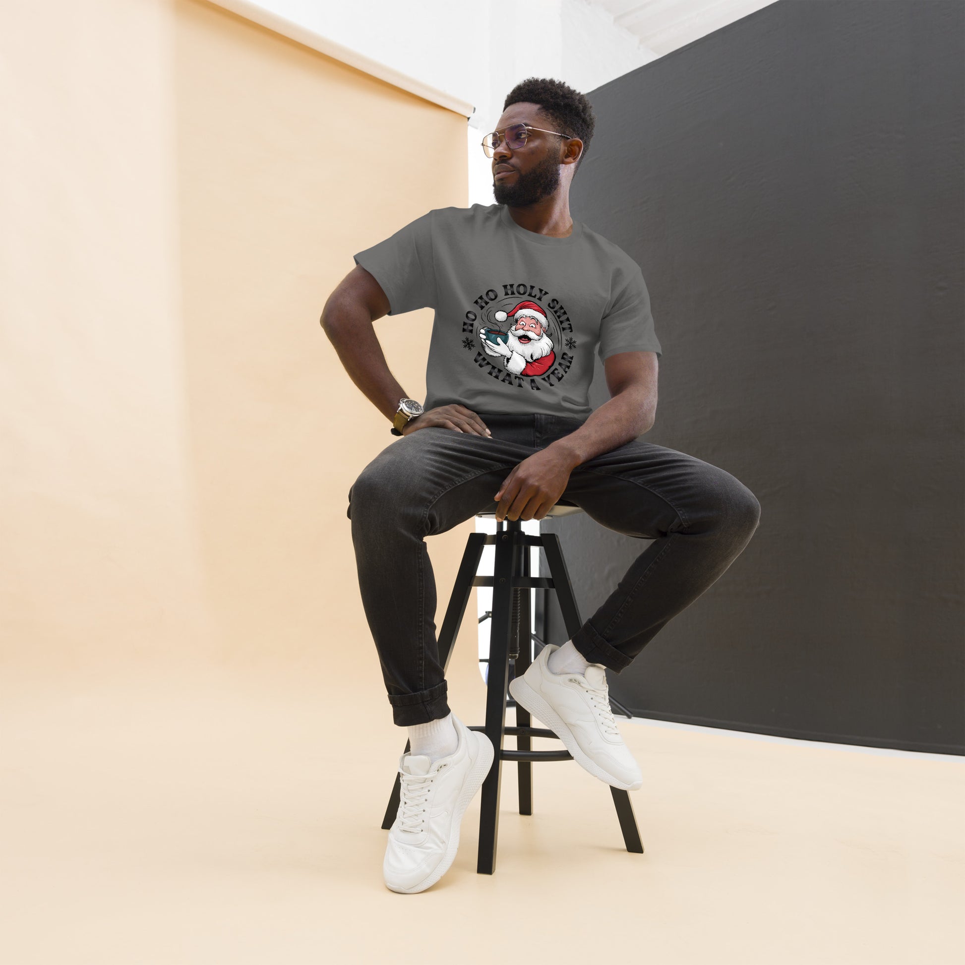 Man sitting on a stool wearing a gray t-shirt with a graphic design, black pants, and white sneakers.
