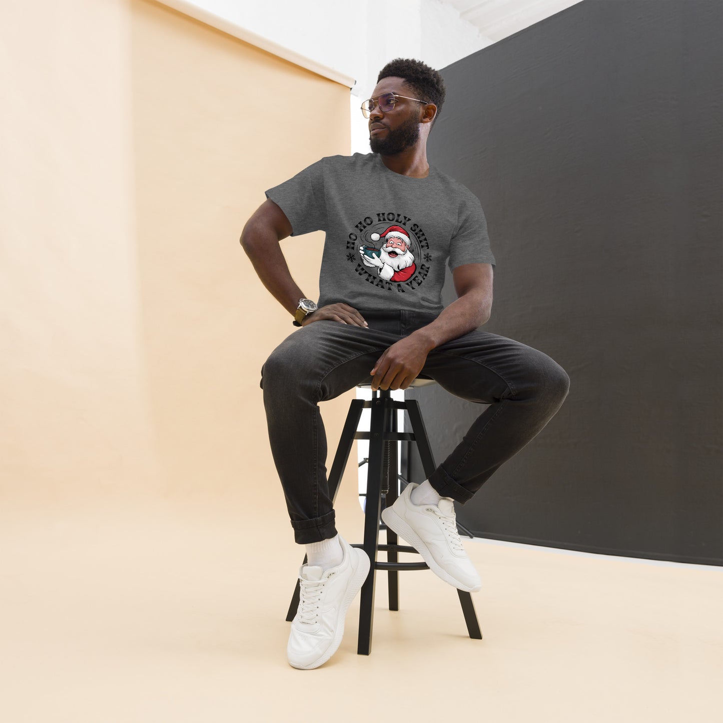 Man sitting on a stool wearing a gray t-shirt with a graphic design, black pants, and white sneakers.