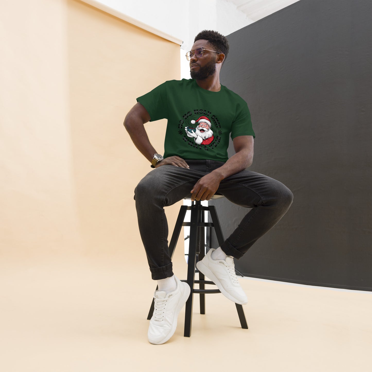 Man sitting on a stool wearing a green t-shirt with a graphic design, black pants, and white sneakers.