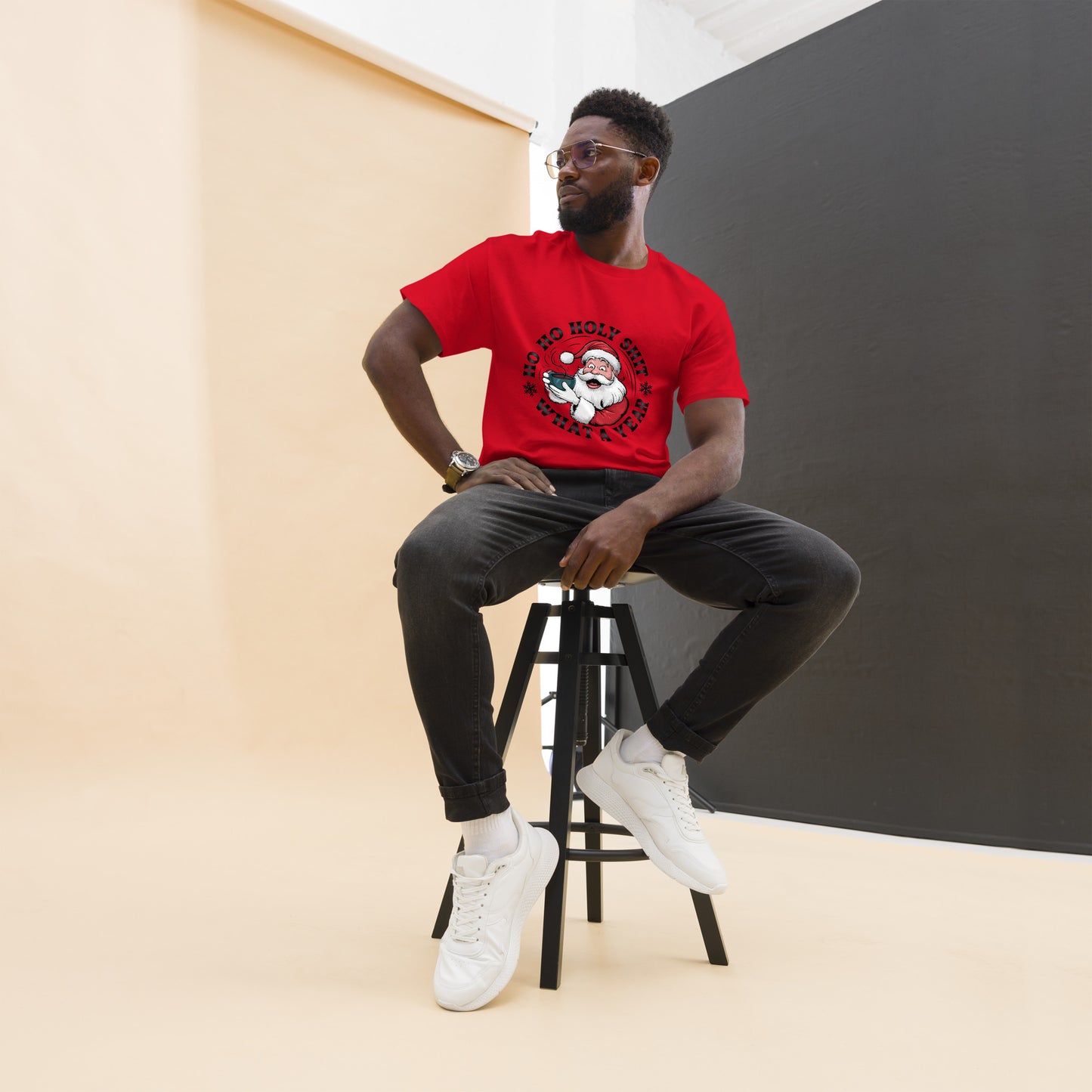 Man sitting on a stool wearing a red t-shirt with a graphic design, black pants, and white sneakers.